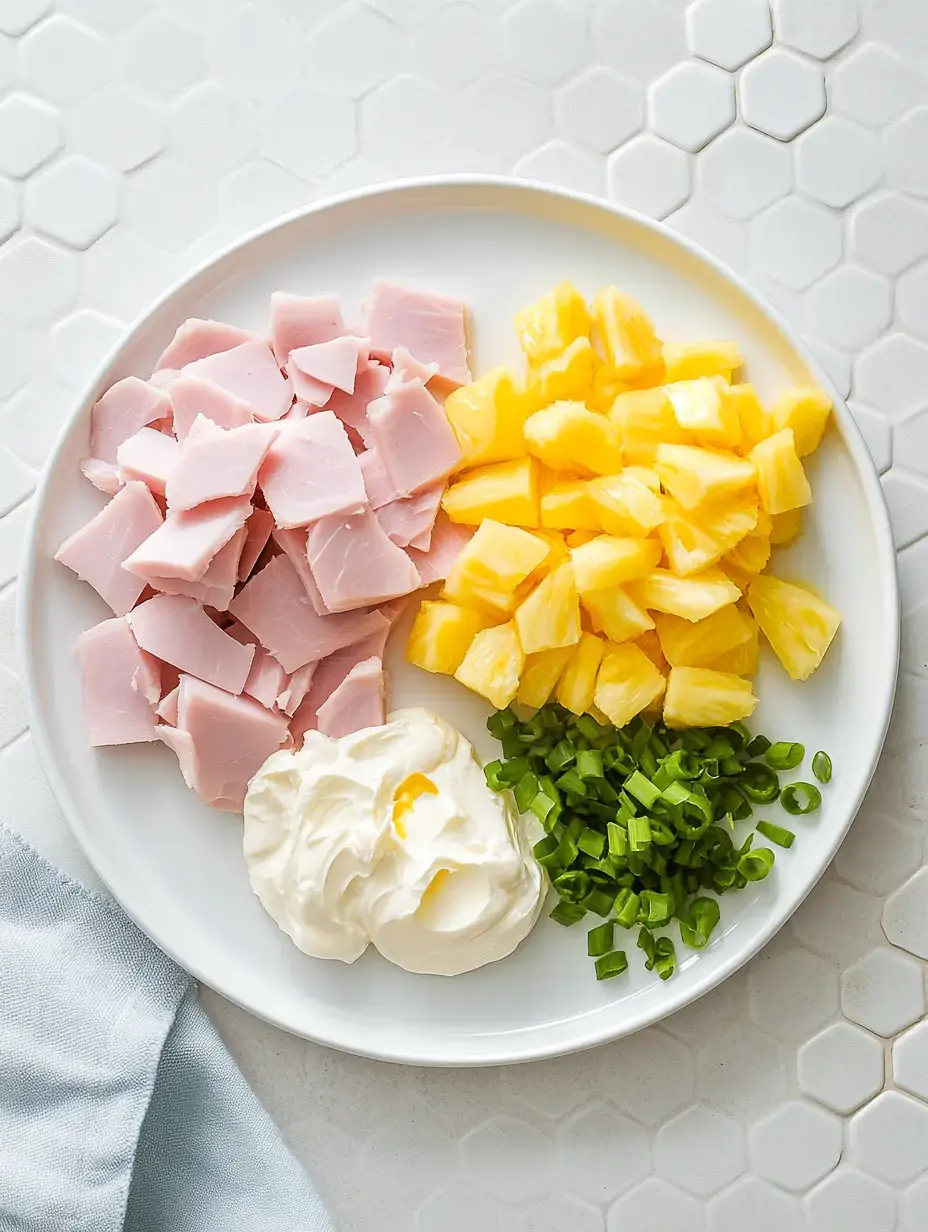 A serving dish showing chopped ham chunks, pineapple pieces, sliced green onions and a cream dollop on a textured surface.