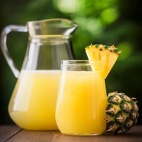 A tall glass of bright yellow pineapple juice next to a bigger pitcher, a whole pineapple, and a soft-focus green background.